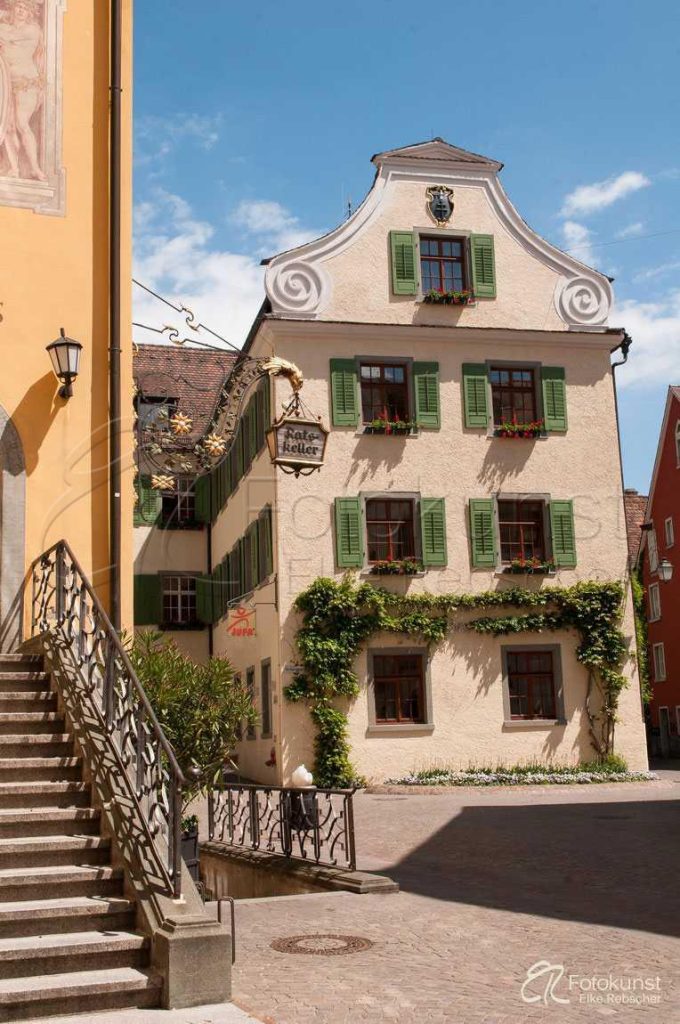 ein Teil des Treppenaufgangs zum gelblich gestrichenen Rathaus der Stadt Meersburg am Bodensee mit einem hellen Geb&auml;ude mit gr&uuml;nen Schlagl&auml;den
